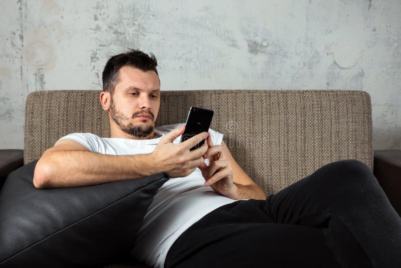 Der Kerl im wei?en Hemd liegt auf der Couch und sitzt im Telefon Das Konzept der Tr?gheit, Apathie lizenzfreie stockbilder Der Kerl im wei?en Hemd liegt auf der Couch und sitzt im Telefon Das Konzept der Tr?gheit, Apathie lizenzfreie stockbilder