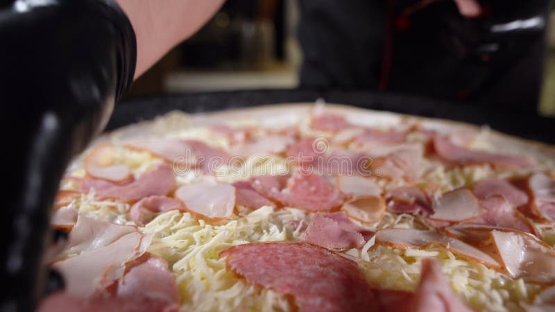 Der Prozess der Herstellung einer großen Pizza. die Hände des Chefs in schwarzen Handschuhen legen den Schinken auf den geriebenen