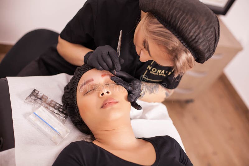 Dermatologista fêmea que usa a máquina das agulhas para a remoção da tatuagem da sobrancelha fotografia de stock royalty free Dermatologista fêmea que usa a máquina das agulhas para a remoção da tatuagem da sobrancelha fotografia de stock royalty free