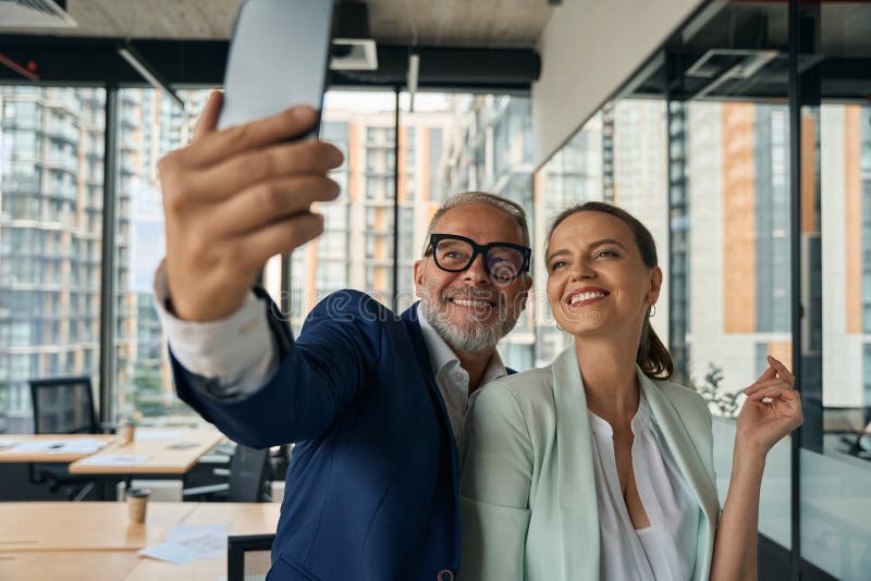 Des collègues joyeux qui prennent le selfie dans un bureau moderne. Happy business man tient un smartphone et utilise l'appareil photo pour attraper un moment avec une collegue féminine sur le lieu de travail