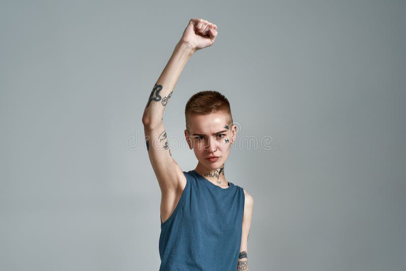 Descontento tatuado joven mujer caucásica manifestante con piercing mirando a cámara mientras posa con el brazo levantado