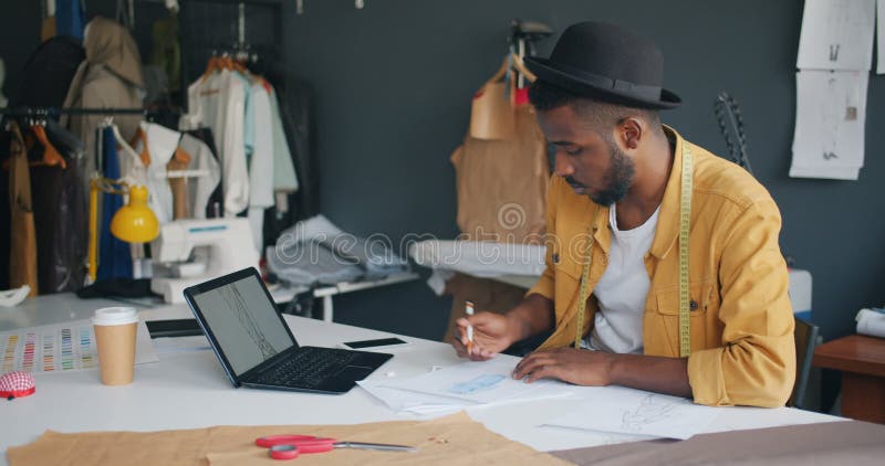 Desenhista de roupa que esboça olhando a tela do portátil que trabalha apenas no estúdio