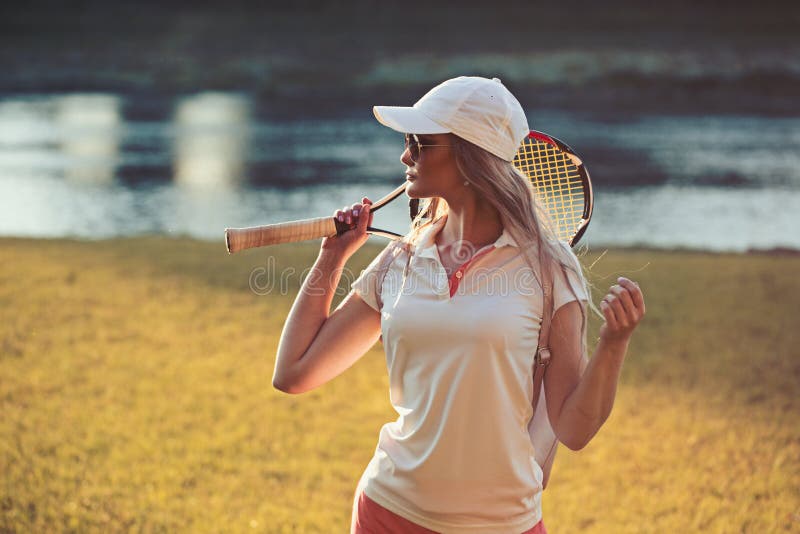 Desportista com a raquete de tênis no rio Mulher da forma no equipamento do tênis na paisagem do verão Mulher no tampão em ensola imagem de stock royalty free Desportista com a raquete de tênis no rio Mulher da forma no equipamento do tênis na paisagem do verão Mulher no tampão em ensola imagem de stock royalty free
