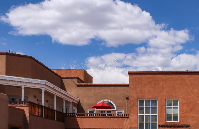 The Rooftops of Santa Fe New Mexico Looking across the street from on rooftop to stucco buildings with balconies and a bright red umbrella with birds sitting on roofline under blue cloudy sky. The Rooftops of Santa Fe New Mexico Looking across the street from on rooftop to stucco buildings with balconies and a bright red umbrella with birds sitting on roofline under blue cloudy sky
