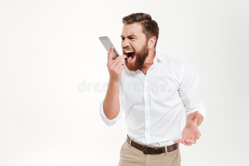 Image of screaming angry young bearded emotional man standing over white wall background isolated. Looking aside while talking by phone. Image of screaming angry young bearded emotional man standing over white wall background isolated. Looking aside while talking by phone.