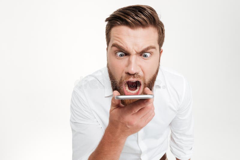Image of screaming angry young bearded emotional man standing over white wall background isolated. Looking aside while talking by phone. Image of screaming angry young bearded emotional man standing over white wall background isolated. Looking aside while talking by phone.