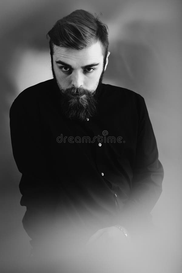 Det svartvita fotoet av en stilfull man med ett iklädda skägg och stilfull frisyr den svarta skjortan står i dimman arkivfoto Det svartvita fotoet av en stilfull man med ett iklädda skägg och stilfull frisyr den svarta skjortan står i dimman arkivfoto