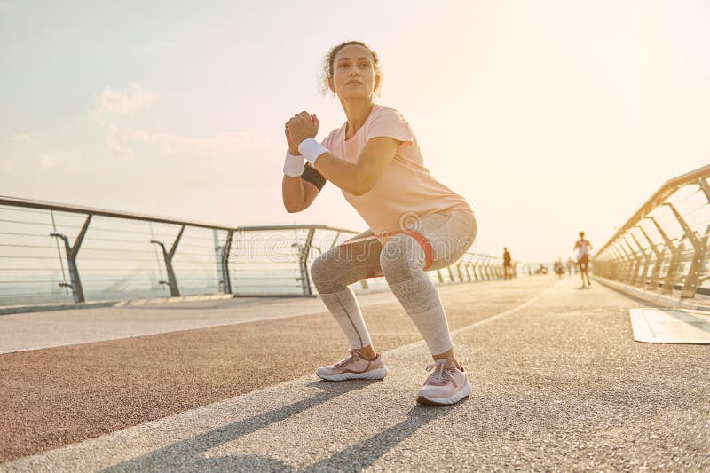 Determined Sporty Woman Exercising Outdoor At Sunrise Doing Squats With Elastic Resistance Band