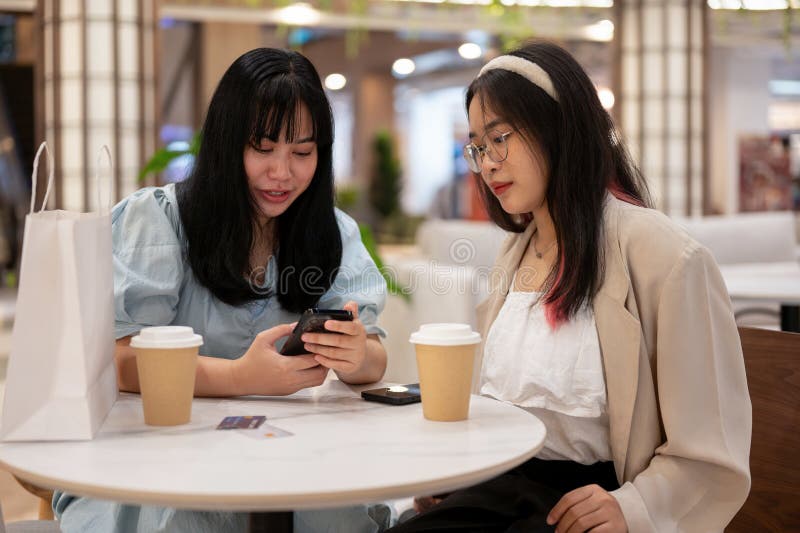 Two young Asian female friends are enjoying talking and looking at something on a smartphone while sitting together in a cafe in the shopping mall. city life and lifestyle concepts. Two young Asian female friends are enjoying talking and looking at something on a smartphone while sitting together in a cafe in the shopping mall. city life and lifestyle concepts