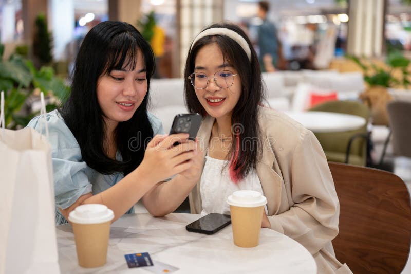 Two young Asian female friends are enjoying talking and looking at something on a smartphone while sitting together in a cafe in the shopping mall. city life and lifestyle concepts. Two young Asian female friends are enjoying talking and looking at something on a smartphone while sitting together in a cafe in the shopping mall. city life and lifestyle concepts