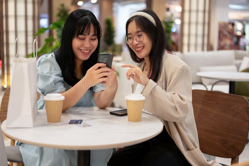 Two young Asian female friends are enjoying talking and looking at something on a smartphone while sitting together in a cafe in the shopping mall. city life and lifestyle concepts. Two young Asian female friends are enjoying talking and looking at something on a smartphone while sitting together in a cafe in the shopping mall. city life and lifestyle concepts