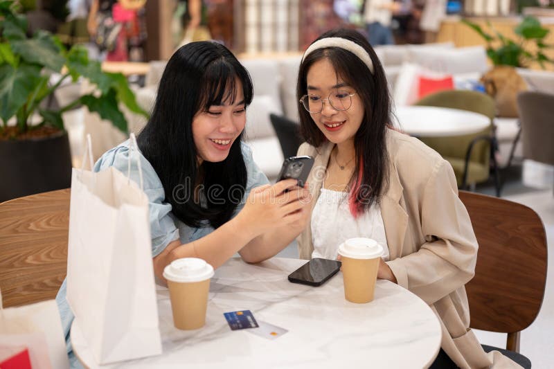 Two young Asian female friends are enjoying talking and looking at something on a smartphone while sitting together in a cafe in the shopping mall. city life and lifestyle concepts. Two young Asian female friends are enjoying talking and looking at something on a smartphone while sitting together in a cafe in the shopping mall. city life and lifestyle concepts