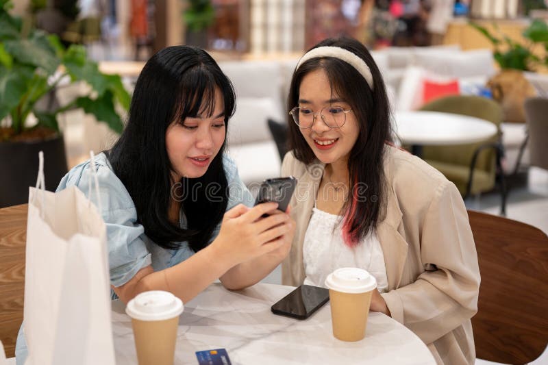 Two young Asian female friends are enjoying talking and looking at something on a smartphone while sitting together in a cafe in the shopping mall. city life and lifestyle concepts. Two young Asian female friends are enjoying talking and looking at something on a smartphone while sitting together in a cafe in the shopping mall. city life and lifestyle concepts