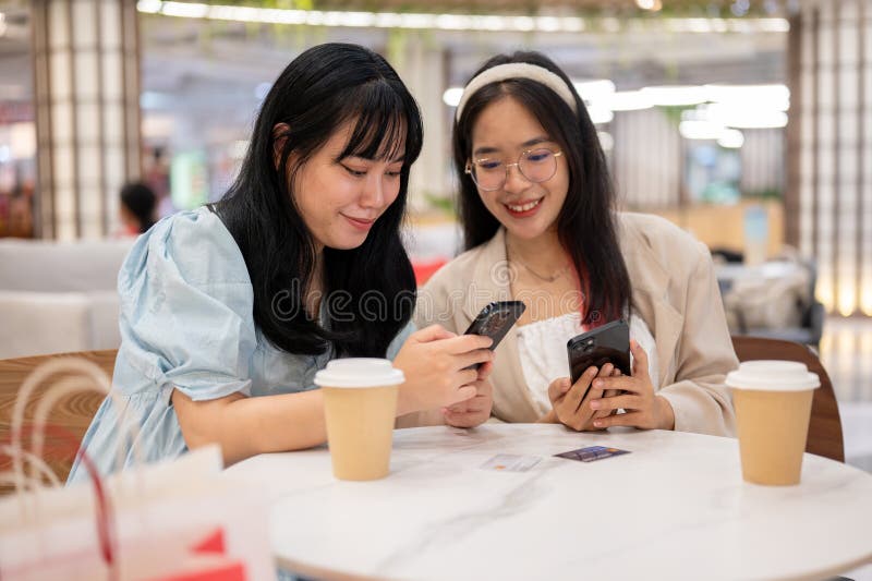 Two cheerful young Asian female friends are enjoying talking about something on their phones while relaxing in a cafe in the shopping mall after shopping together. lifestyle and friendship concepts. Two cheerful young Asian female friends are enjoying talking about something on their phones while relaxing in a cafe in the shopping mall after shopping together. lifestyle and friendship concepts