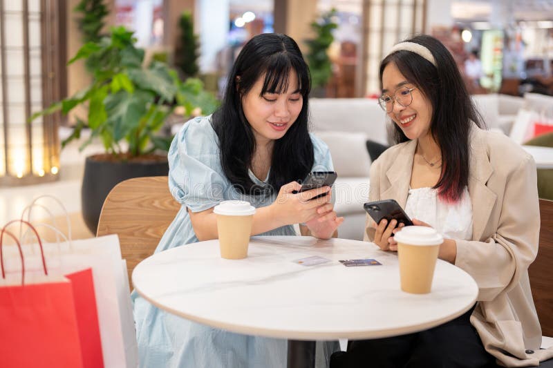 Two cheerful young Asian female friends are enjoying talking about something on their phones while relaxing in a cafe in the shopping mall after shopping together. lifestyle and friendship concepts. Two cheerful young Asian female friends are enjoying talking about something on their phones while relaxing in a cafe in the shopping mall after shopping together. lifestyle and friendship concepts