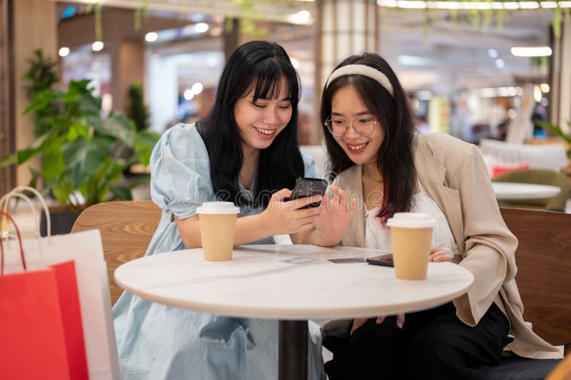 Two cheerful young Asian female friends are enjoying talking about something on their phones while relaxing in a cafe in the shopping mall after shopping together. lifestyle and friendship concepts. Two cheerful young Asian female friends are enjoying talking about something on their phones while relaxing in a cafe in the shopping mall after shopping together. lifestyle and friendship concepts
