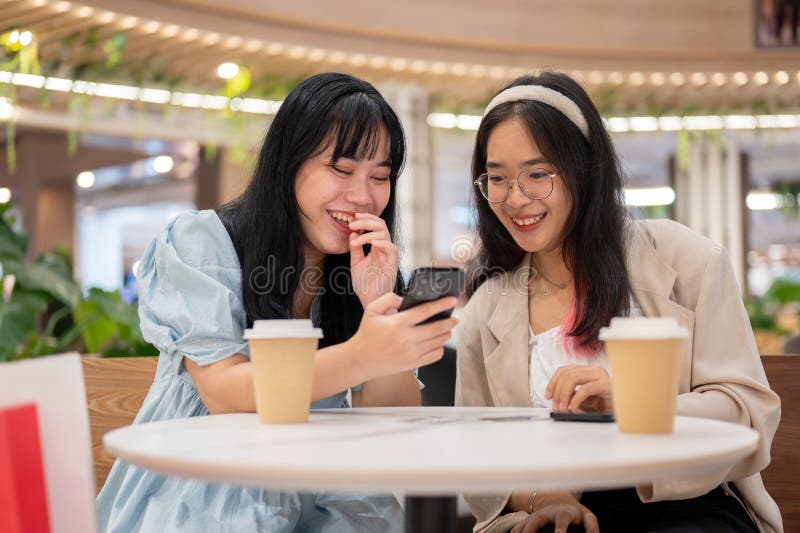 Two cheerful young Asian female friends are enjoying talking about something on their phones while relaxing in a cafe in the shopping mall after shopping together. lifestyle and friendship concepts. Two cheerful young Asian female friends are enjoying talking about something on their phones while relaxing in a cafe in the shopping mall after shopping together. lifestyle and friendship concepts