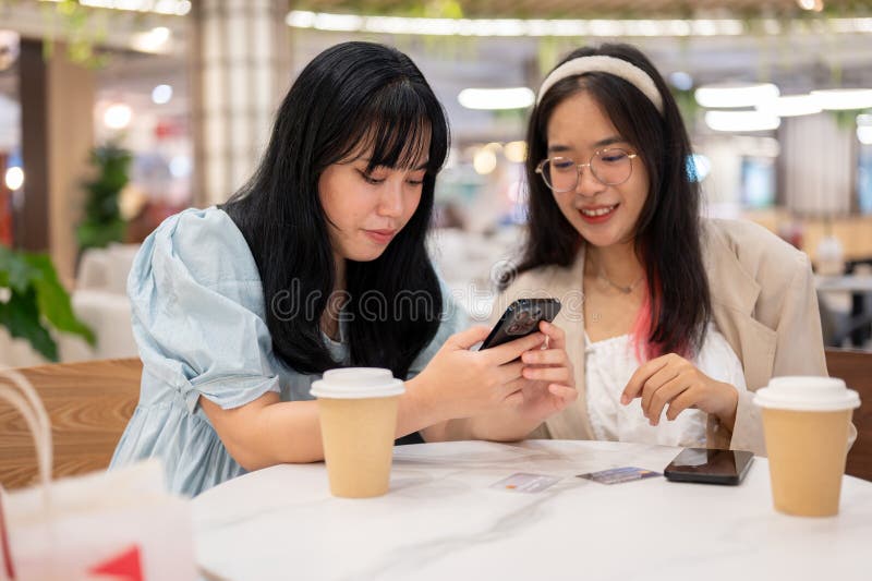 Two cheerful young Asian female friends are enjoying talking about something on their phones while relaxing in a cafe in the shopping mall after shopping together. lifestyle and friendship concepts. Two cheerful young Asian female friends are enjoying talking about something on their phones while relaxing in a cafe in the shopping mall after shopping together. lifestyle and friendship concepts