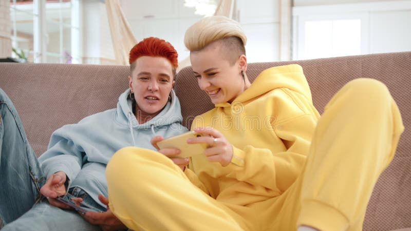 Deux amis qui s'amusent à jouer sur des appareils mobiles à l'intérieur rient et interagissent sur un confortable canapé banque de vidéos