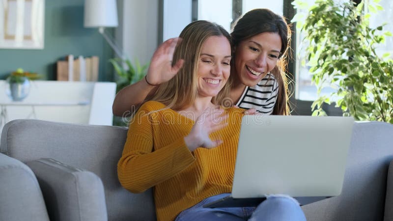 Deux belles femmes heureuses faisant un appel vidéo avec ordinateur portable tout en s'embrassant assis sur le canapé à la maison. banque de vidéos