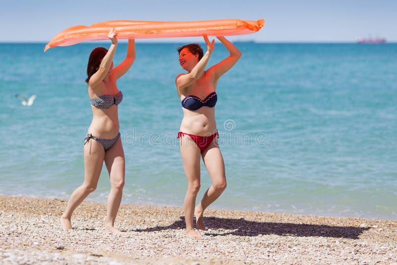 Two girls at the sea. Women in swimsuits carrying inflatable raft over their heads walking along beach. Two girls at the sea. Women in swimsuits carrying inflatable raft over their heads walking along beach