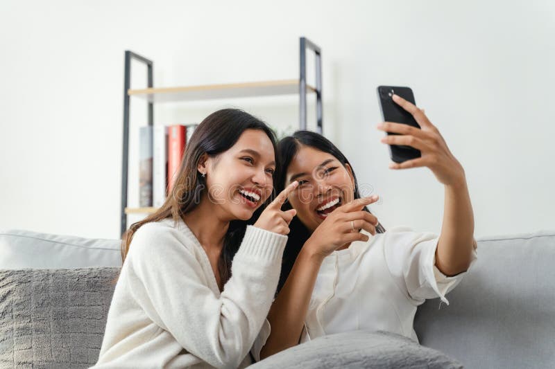 Two women are sitting on a couch and taking a selfie. They are both smiling, laughing and pointing at the Smart phone screen. Two women are sitting on a couch and taking a selfie. They are both smiling, laughing and pointing at the Smart phone screen
