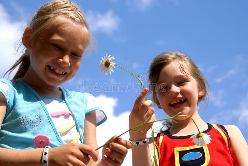 Two laughing girls hold in camomile hands flowers - camomiles against the sky. Two laughing girls hold in camomile hands flowers - camomiles against the sky