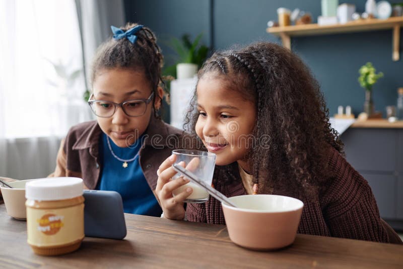 Portrait of two African American girls watching videos via smartphone at kitchen table during breakfast. Portrait of two African American girls watching videos via smartphone at kitchen table during breakfast