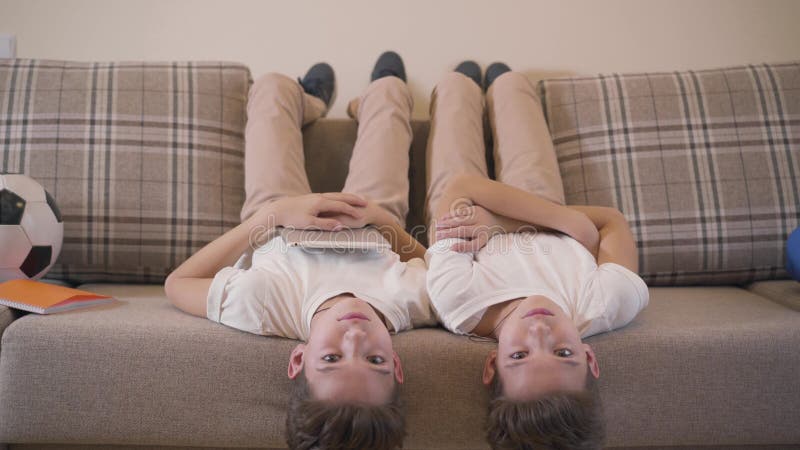 Deux frères jumeaux caucasiens allongés sur un canapé, regardant la caméra et riant Les frères et soeurs joyeux qui reposent banque de vidéos