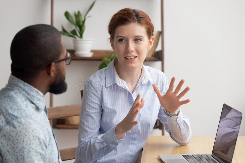 Two diverse businesspeople chatting sitting behind laptop in office. Excited caucasian female sharing ideas or startup business plan with black male coworker. Informal conversation, work break concept. Two diverse businesspeople chatting sitting behind laptop in office. Excited caucasian female sharing ideas or startup business plan with black male coworker. Informal conversation, work break concept