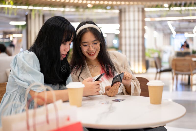 Two happy young Asian female friends are hanging out together at the shopping mall on the weekend, talking about something on their phones while sitting in a cafe. lifestyle concept. Two happy young Asian female friends are hanging out together at the shopping mall on the weekend, talking about something on their phones while sitting in a cafe. lifestyle concept