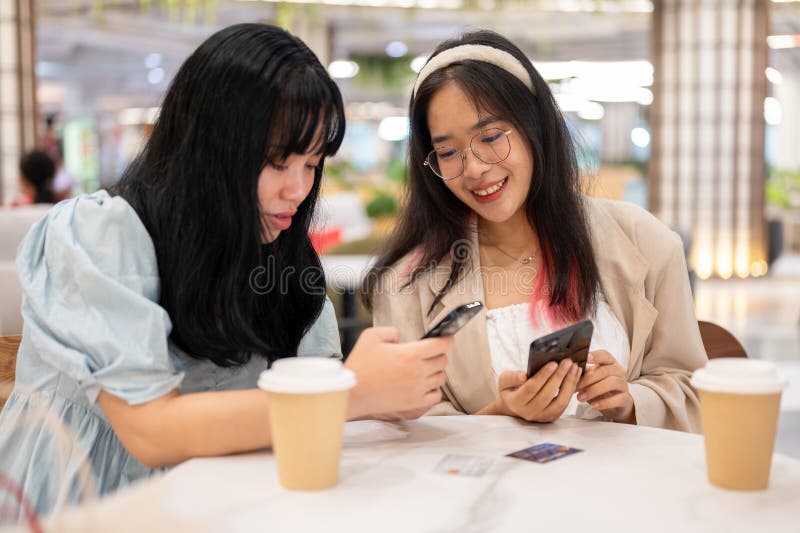 Two happy young Asian female friends are hanging out together at the shopping mall on the weekend, talking about something on their phones while sitting in a cafe. lifestyle concept. Two happy young Asian female friends are hanging out together at the shopping mall on the weekend, talking about something on their phones while sitting in a cafe. lifestyle concept