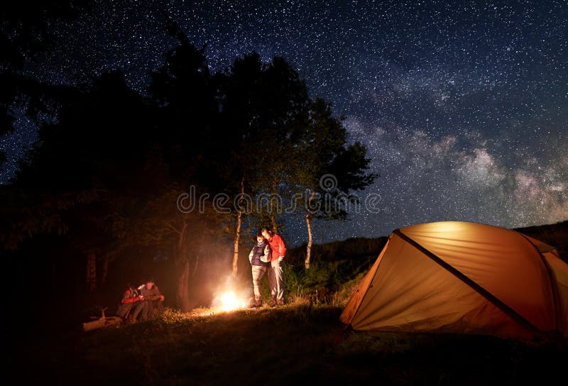 Deux jeunes couples au feu de camp sous le ciel étoilé lumineux près de la tente sur le fond des arbres images stock Deux jeunes couples au feu de camp sous le ciel étoilé lumineux près de la tente sur le fond des arbres images stock
