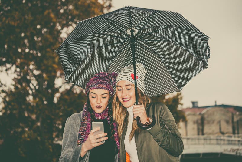Deux jeunes femmes avec le parapluie et le téléphone portable d'utilisation dans le stre photo stock Deux jeunes femmes avec le parapluie et le téléphone portable d'utilisation dans le stre photo stock