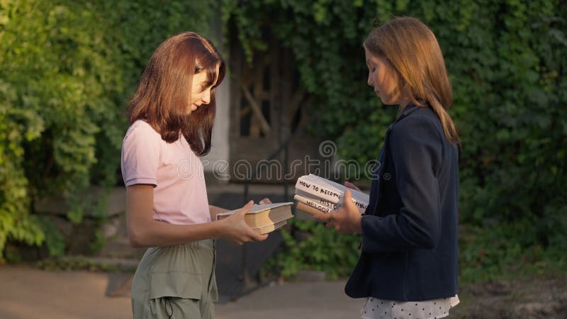 Deux jeunes filles intelligentes échangeant des livres au ralenti au coucher du soleil au parc d'été du printemps. vue latérale po clips vidéos