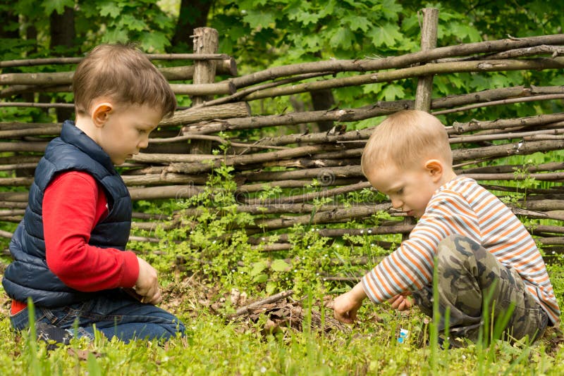 Two young boys playing at lighting a campfire kneeling down together carefully applying a lit match to a pile of twigs and wood that they have gathered at a rustic campsite. Two young boys playing at lighting a campfire kneeling down together carefully applying a lit match to a pile of twigs and wood that they have gathered at a rustic campsite