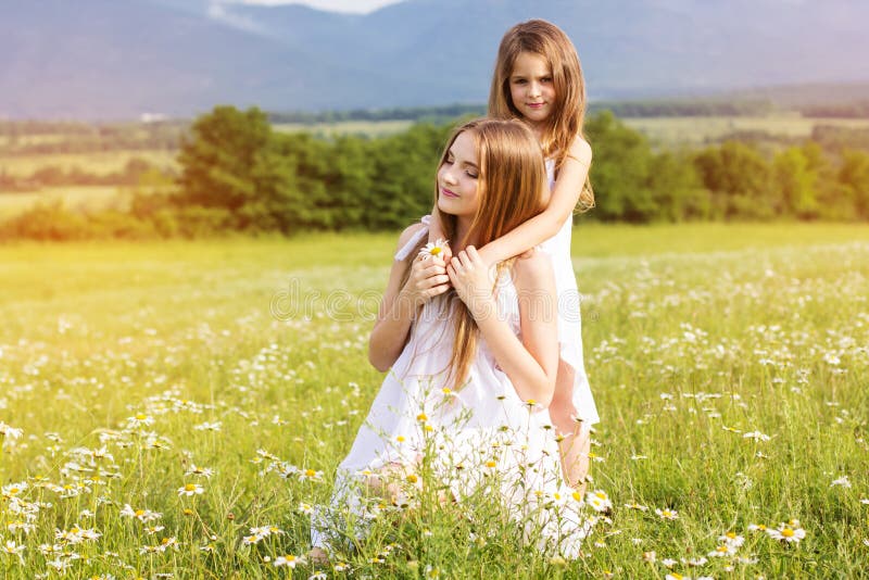 Cute sisters girls are wearing white dresses at green camomile field with mountains view, sunset time. Cute sisters girls are wearing white dresses at green camomile field with mountains view, sunset time