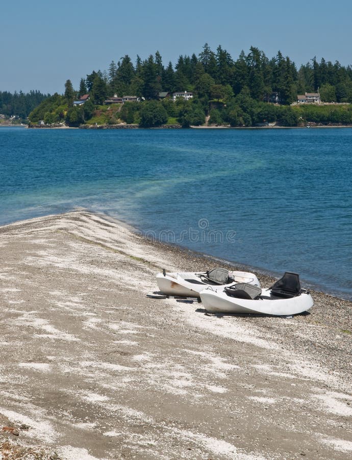 Two kayaks on a sandy spit that trails into the tidal water and points to the distance shore that has large houses and docks among the trees near Gig Harbor, Washington. Two kayaks on a sandy spit that trails into the tidal water and points to the distance shore that has large houses and docks among the trees near Gig Harbor, Washington.