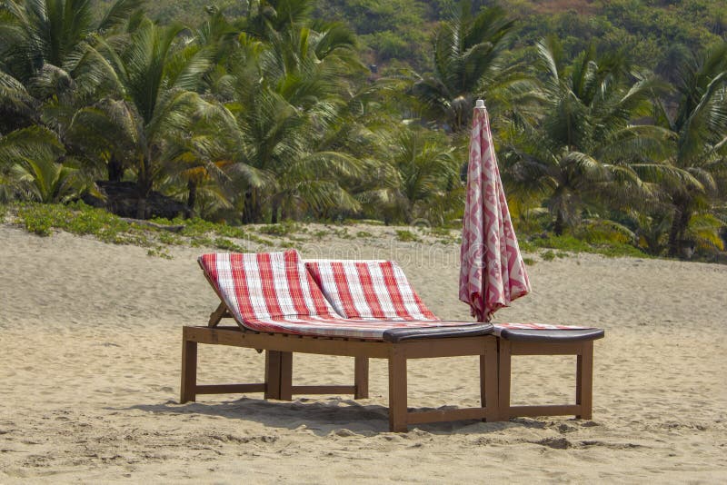 Deux lits en bois de plage avec des matelas rouges et un parapluie plié sur le sable contre le contexte d'une jungle verte troubl photos stock Deux lits en bois de plage avec des matelas rouges et un parapluie plié sur le sable contre le contexte d'une jungle verte troubl photos stock
