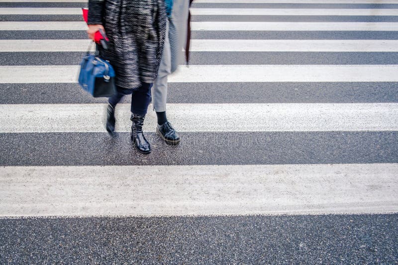 two unrecognizable persons in black shoes cross wet street after rain on crosswalk, red umbrella, parallel lines of pedestrian crossing. two unrecognizable persons in black shoes cross wet street after rain on crosswalk, red umbrella, parallel lines of pedestrian crossing