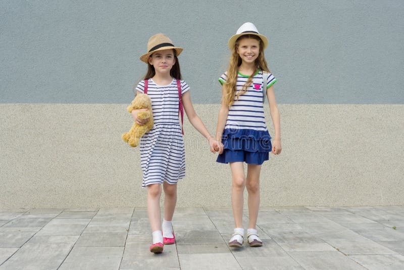 Deux petites belles amies tenant des mains, filles marchant dans des robes rayées, chapeaux avec le sac à dos photographie stock Deux petites belles amies tenant des mains, filles marchant dans des robes rayées, chapeaux avec le sac à dos photographie stock