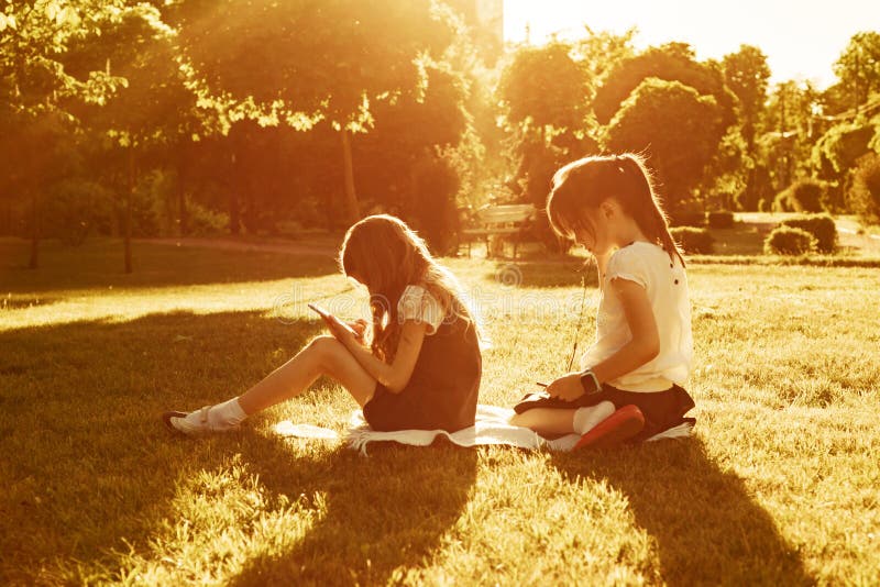 Deux petites écolières à l'aide d'un smartphone Enfants jouant, lecture, regardant le téléphone, en parc, heure d'or Les gens, images stock Deux petites écolières à l'aide d'un smartphone Enfants jouant, lecture, regardant le téléphone, en parc, heure d'or Les gens, images stock