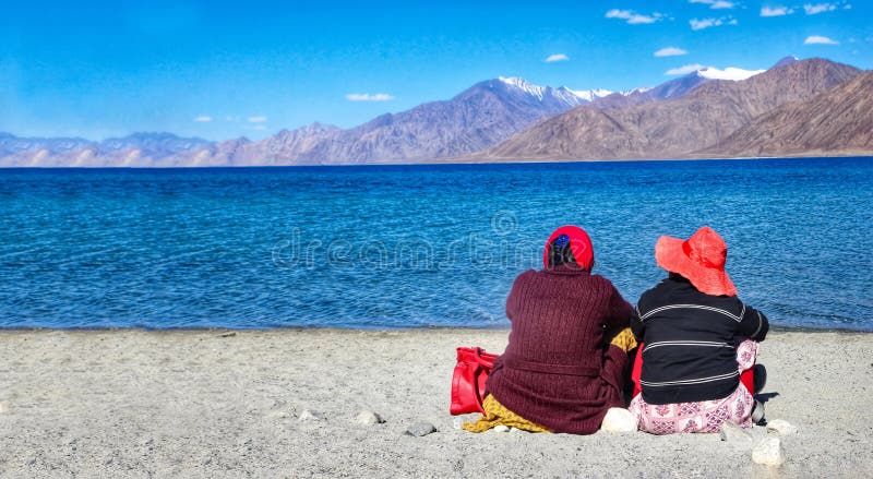 Deux touristes s'asseyant près du lac dans la solitude au cours de la journée devant les eaux bleues et les montagnes éloignées photographie stock Deux touristes s'asseyant près du lac dans la solitude au cours de la journée devant les eaux bleues et les montagnes éloignées photographie stock