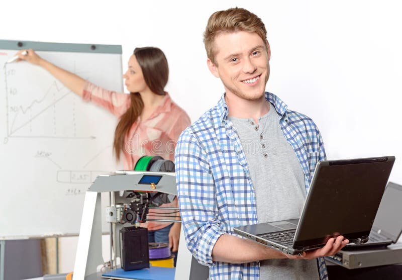 Young handsome science student smiling and holding a laptop in his hands while his colleague drawing diagram on a flip chart board in a laboratory, selective focus. Young handsome science student smiling and holding a laptop in his hands while his colleague drawing diagram on a flip chart board in a laboratory, selective focus