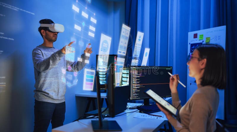 Developer uses a VR headset to interact with projected interface designs, while colleagues discuss and code on screens at office royalty free stock image