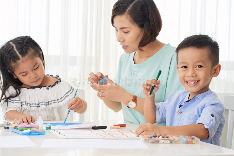Dibujo feliz de los niños y de los estudiantes. Niño pequeño asiático que mira la cámara y que sonríe mientras que dibuja con el profesor en clase de arte