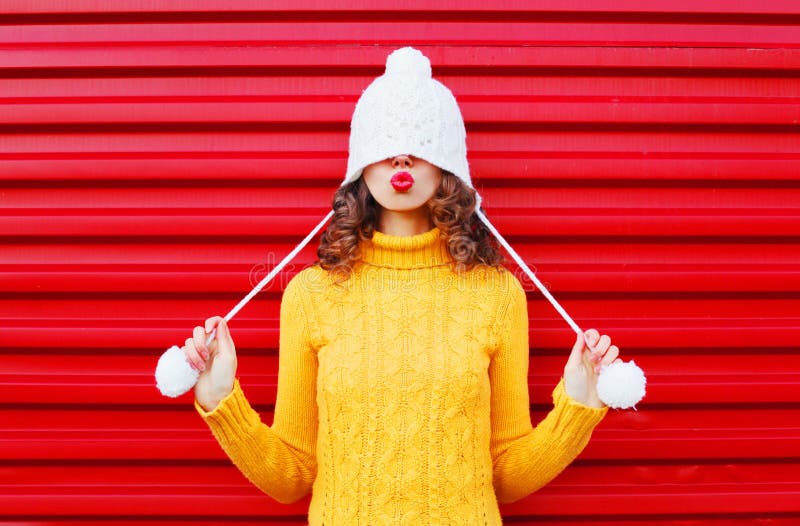 Die glückliche Frau der Mode, die rote Lippen durchbrennt, lässt Luft tragende bunte Strickmütze, gelbe Strickjacke küssen über R stockfotos
