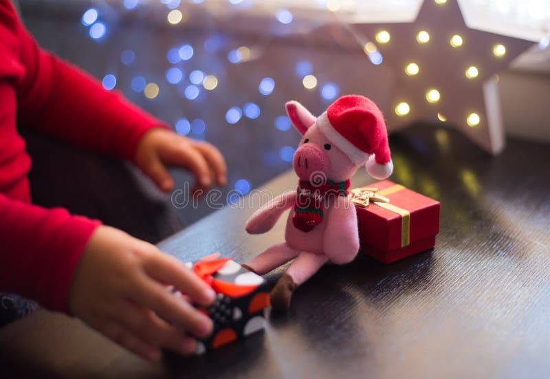 Die Hände der Kinder, die zu Hause Geschenkbox nahe Spielzeugschwein in Sankt-Hut mit Girlande bokeh Hintergrund Innen halten lizenzfreie stockfotografie Die Hände der Kinder, die zu Hause Geschenkbox nahe Spielzeugschwein in Sankt-Hut mit Girlande bokeh Hintergrund Innen halten lizenzfreie stockfotografie