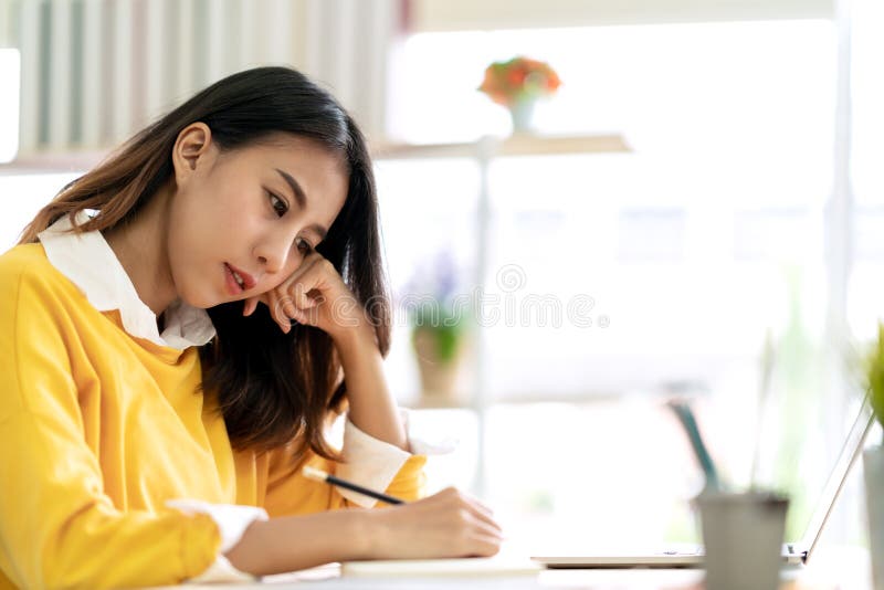 Die junge attraktive asiatische Studentin, die bei Tisch das Denken und das Schreiben der Zeitschrift merken sitzt eigenhändig, I lizenzfreie stockfotografie
