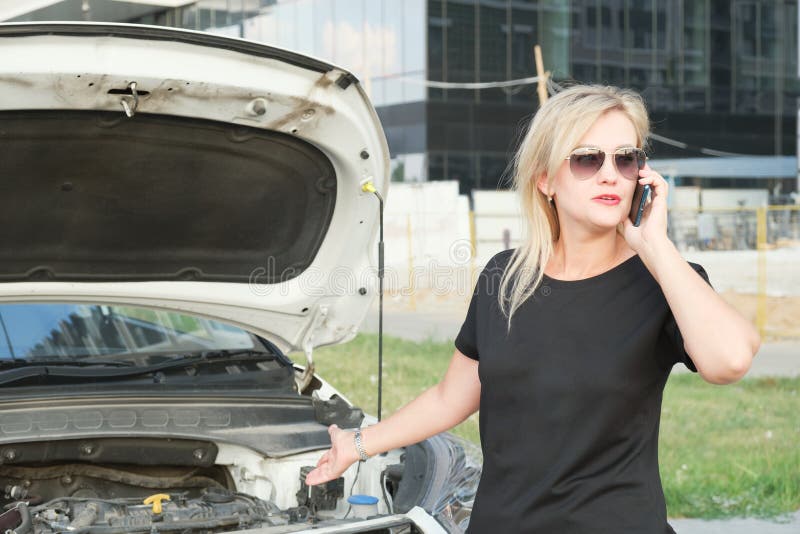 Young woman is standing in front of her car with the hood up and talking on the phone. A woman calls a technical service because of a broken car. The woman is holding her head. Young woman is standing in front of her car with the hood up and talking on the phone. A woman calls a technical service because of a broken car. The woman is holding her head